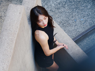 Graceful young woman with long black hair sit casually and lean against the wall, looking away emotionally. Outdoor close up photo of cool Chinese girl in trendy summer sexy black dress. 