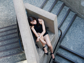 Graceful young woman with long black hair sit casually and lean against the wall, looking away emotionally. Outdoor close up photo of cool Chinese girl in trendy summer sexy black dress. 