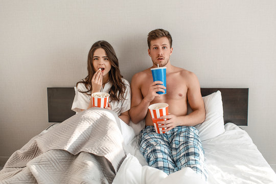 Young couple in the bed. An intrigued beautiful man and woman are eating popcorn and watching TV together in bedroom