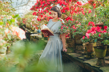 Beautiful adult girl in an azalea greenhouse reading a book and dreaming in a   beautiful retro dress