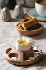 Breakfast with cup of coffee and soft boiled egg, served in wooden egg cup with salt, pepper and toasted bread, jug of cream over linen tablecloth.