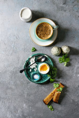 Breakfast with cup of coffee and soft boiled egg, served in green ceramic egg cup with salt, pepper and toasted bread, jug of cream over grey texture background. Flat lay, space