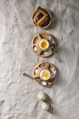 Breakfast with soft boiled eggs, served in wooden egg cup with salt, pepper and toasted bread in row over linen tablecloth. Flat lay, space