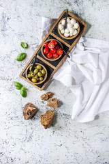 Mozzarella, cherry tomatoes, olives antipasto appetizers served in wooden bowl on slate serving board with basil and rye bread over white marble background. Flat lay, space