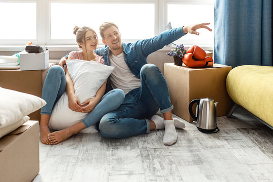 Young family couple bought or rented their first small apartment. People planning and imagine. They sit on floor. Guy hugs young woman and point. Moving in and unpacking.