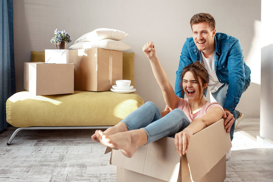 Young family couple bought or rented their first small apartment. Cheerful woman scream sitting in box. Guy move her. They play game during moving in.