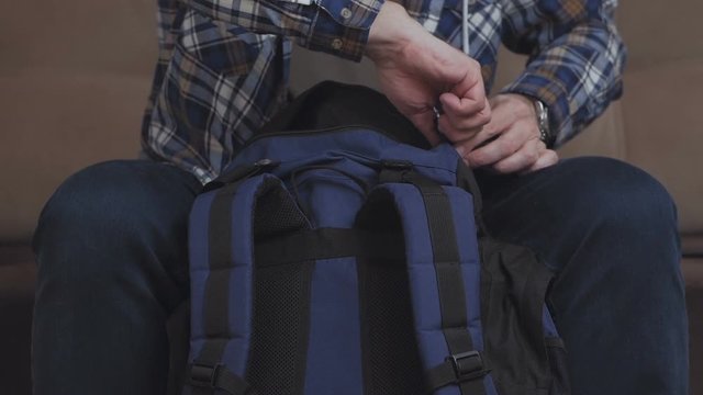 A man sits on the couch, puts things in a backpack, fastens it and leaves the frame.