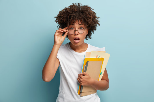 Embarrssed Emotional Lady With Dark Skin, Carries Spiral Notebook, Papers, Touches Frame Of Glasses, Looks Surprisingly At Timetable In University, Wonders Some Changes, Wears White T Shirt.