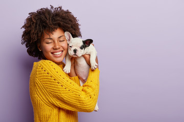 Charming happy woman of Afro appearance expresses positive emotions during photoshoot with french bulldog puppy, cuddles gently near face, smiles broadly, laughs at camera, blank space aside