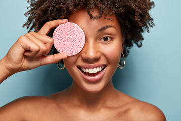 Positive lady stands with naked shoulders, holds cosmetic sponge over eye, smiles happily, has...