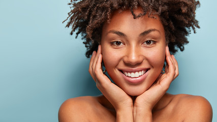 Close up portrait of relaxed black woman has gentle skin after taking shower, satisfied with new lotion, has no makeup, smiles tenderly, shows perfect teeth, stands shirtless against blue background - obrazy, fototapety, plakaty