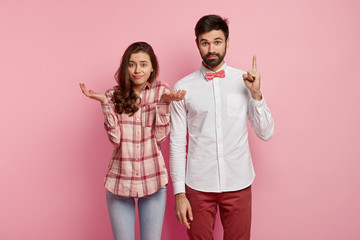 Hesitant clueless woman shruggs shoulders, surprised unshaven man has idea, raises fore finger, collaborate together, pose indoor against pink background. Couple or friends find right decision