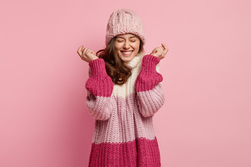 Isolated shot of overjoyed woman raises hands, wears knitted hat and loose sweater, expresses...