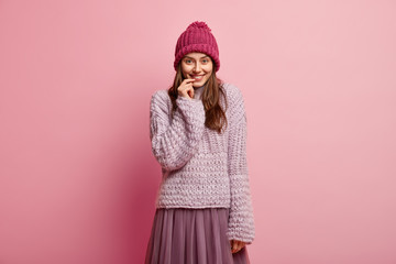 Pretty lady with satisfied expression, keeps hand near lips, hears something positive, wears winter outfit, looks at camera with pleasure and delight, models over rosy background. Emotions concept