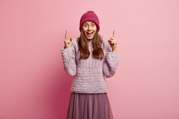 Happy lady expresses surprisement and joy, shows direction upwards, indicates with both fore fingers, dressed in winter clothing, poses against pink background, gestures emotionally, has great idea