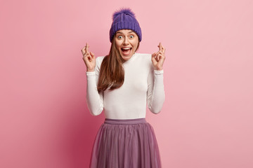 Obraz na płótnie Canvas Positive young woman keeps fingers crossed, exclaims with joy, feels overemotive, believes in good fortune, hopes dreams come true, wears purple hat with pompon, gestures against pink background