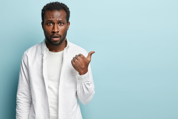 Curly ethnic adult man with indignant expression points aside, has black healthy skin, wears white...