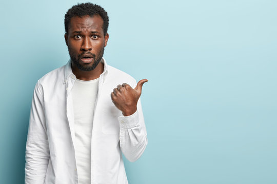 Curly ethnic adult man with indignant expression points aside, has black healthy skin, wears white clothes, puzzled to advertise thing, stands against blue wall, empty space. Damn, what is there.