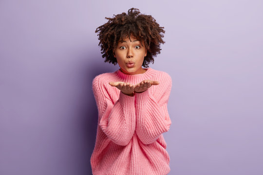 Photo of attractive young woman spreads hands near face, makes mwah to camera, dressed in oversized jumper, expresses love to beloved person, isolated over purple background. Flirting concept