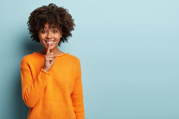 Glad dark skinned young lady makes silence gesture, has toothy smile, tells pleasant secret information to close friend, has friendly expression, stands against blue background with blank space