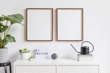 Obraz na płótnie Canvas Minimalistic home decor of interior with two brown wooden mock up photo frames on the white shelf with books, black watering can tropical plants, box and home accessories. White walls. Mockup concept.