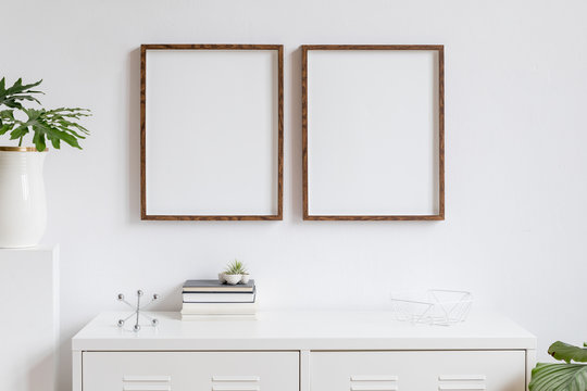 Stylish home interior with two brown wooden mock up photo frames on the white shelf with books, beautiful leaf, stand and home accessories. Minimalistic concept of white room decor.