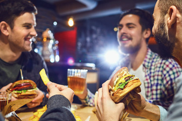 Happy friends eat burgers, drink beer in a bar. - obrazy, fototapety, plakaty