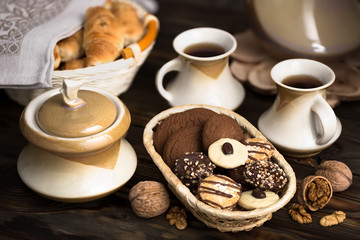 On the table: two cups of tea, sugar bowl and cookies in chocolate glaze sprinkled with caramel crumbs and crushed nuts in a wicker dish.