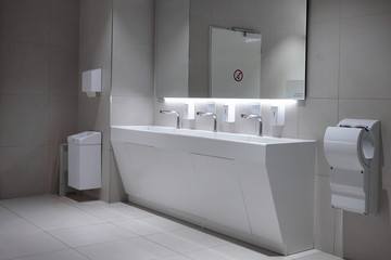 Empty modern ladies restroom with large mirror on gray tiled wall, one sink and three faucets. Public toilet with hand dryer, ceramic basin for washing hands and paper towels, nobody around