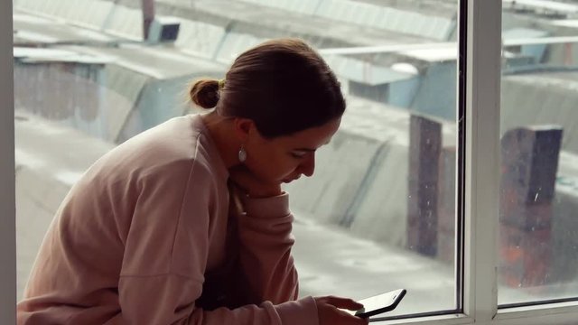 A girl is sitting with a telephone near a large window. She thinks about something and sad. Close-up.