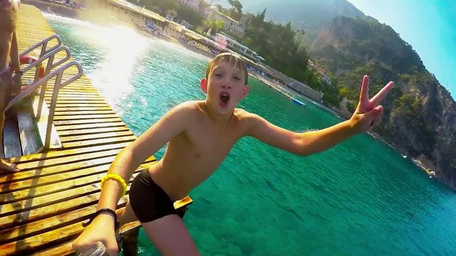 Teenage boy dives into the blue sea from the bridge and makes selfie video.   Happy kid is diving from the wooden bridge into water. Slow motion. Mediterranean sea. Beautiful landscape. Turkey.