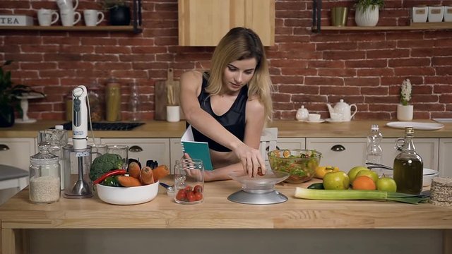 Young sportswoman is weighting cherry tomatoes for prepare healthy food and recording recipe in a notebook at kitchen. Healthy lifestyle, diet