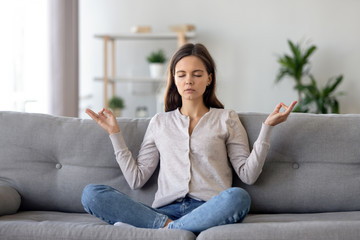 Peaceful young woman sitting in lotus pose on sofa, meditating at home