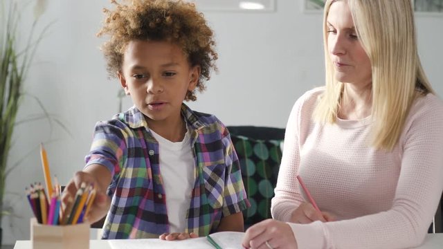 Attractive blond hair woman mom in casual wear helping prepare difficult task at school homework to her lovely little son. They sitting together behind table inside bright light living room and