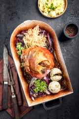 Traditional Bavarian schweinshaxe with onion, potato salad and sauerkraut as top view in a rustic stewpot on an old board