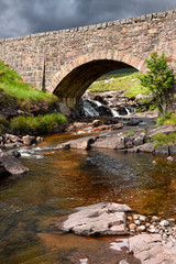 Fototapeta na wymiar Red stone bridge in sun over Russel Burn river of Bealach na Ba road mountain pass on Applecross Peninsula Scottish Highlands Scotland UK