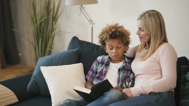 Calm and blond hair woman mama, wearing in casual wear sitting on cozy comfort sofa inside bright light living room. She spending free time with her charming and sweet son, reading interesting books