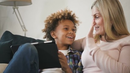 Glad and positive woman mom sitting on comfort couch inside bright light living room with her glad and excited son. They looking at tablet together, making wide beaming smile and clapping in palm - Powered by Adobe
