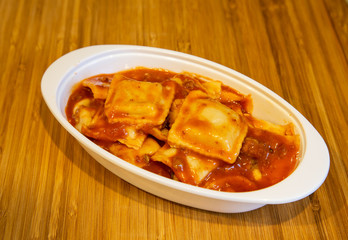 Fresh cooked ravioli in a tomato sauce in a white dish on a wood cutting board.