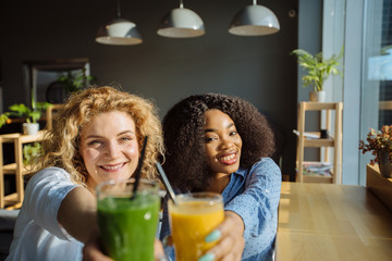 Multi-ethnic curly women friends holding and clink glasses with fresh smoothies from fruits, in modern interior cafe on background. African and american friendship concept.