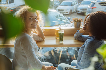 Multi-cultural and difference of cultures, friendship concept. Two curly multiracial female friends talking drinking fresh smoothies or juice in cafe interior over window on backround. Side view