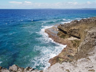 Gentle waves from blue waters slapping against sharp cliffs in a tropical island beach. Seen in the far distance is the island of Tinian, Northern Mariana Islands.