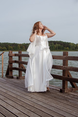 Red-haired bride in a white dress posing on a quay near a lake