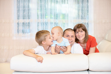 Obraz premium Family concept, young parents with two children, baby bites his brother's nose. Funny, happy childhood. Family on the couch looking at the camera, smiling. a happy family