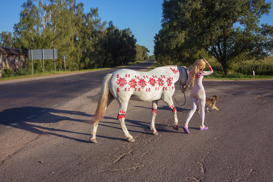 Blonde girl in white bodypainting walking on the road with a white horse