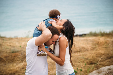Happy young family in white t-shirts and blue jeans with a small daughter in dress kissing near to the lighthouse, playing together