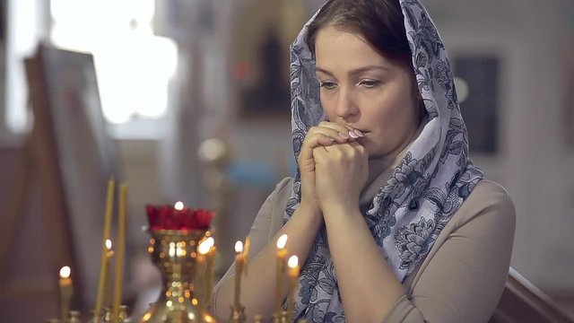 woman in the Russian Orthodox Church with red hair and a scarf on her head lights a candle and prays in front of the icon.