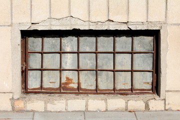 Basement window made of strong frame with metal bars and sheet metal filling mounted on house wall with dilapidated stone tiles locked with rusted padlock on warm sunny day