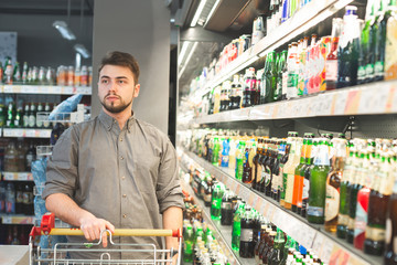 Portrait of a man with a cart, standing in the alcohol department of the supermarket near the shelves with bottles of beer. Male buyer in the beer department at the supermarket. Shopping concept.