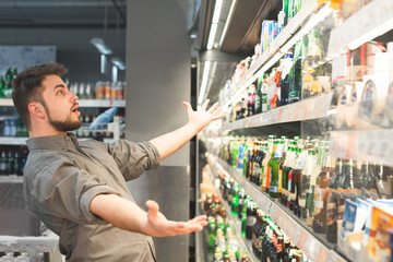 Funny adult looks at the shelf with bottles of beer and can not choose, he wants to take...